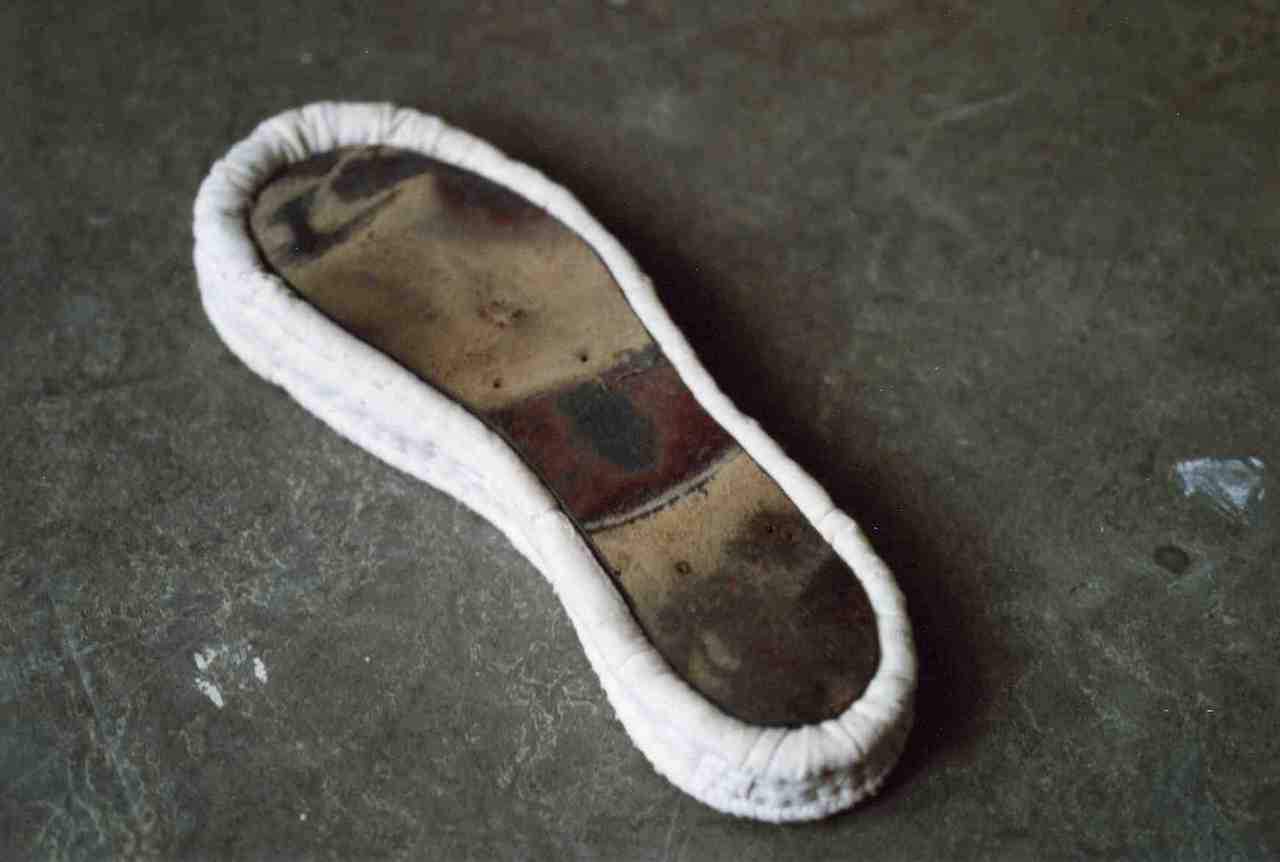 Semelle en tissu évoquant un matelas molletonné en forme de semelle, une semelle en cuir est incrustée dans le matelas, face verso.