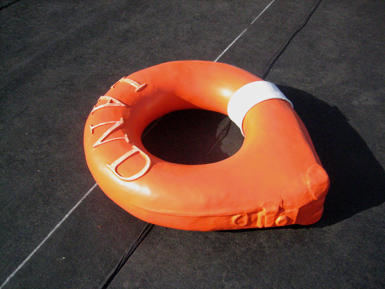 Sculpture évoquant une bouée de sauvetage orange avec un coin de valise moulé et le mot LAND imprimé sur le dessus
