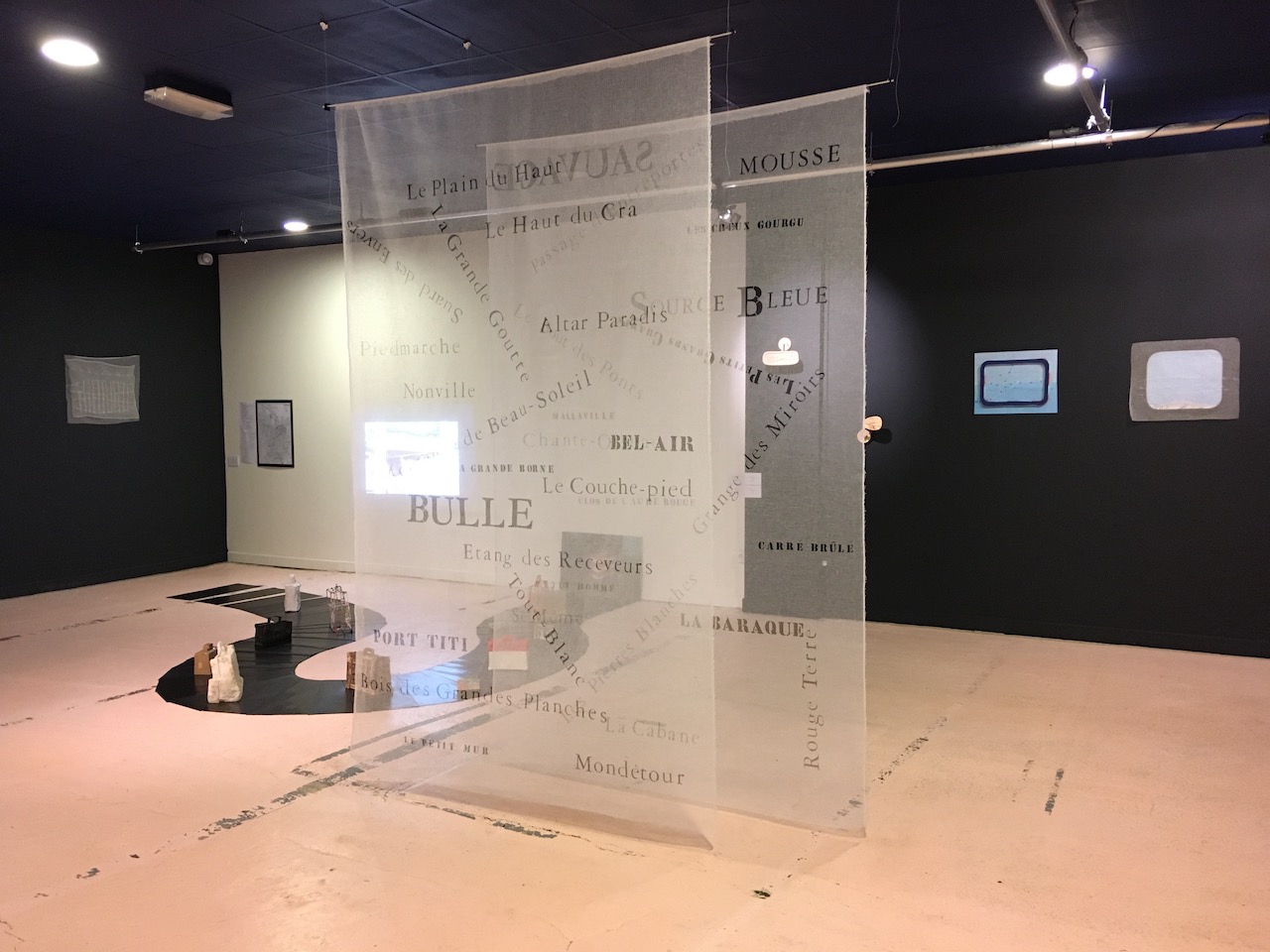 Vue d'exposition, avec une installation de valise sur tapis roulant en caoutchouc que l'on voit à travers des textiles translucides.