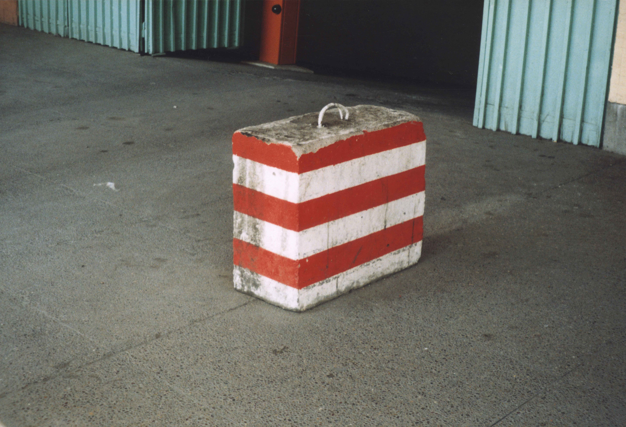 Photo d'un bloc de béton posé dans la rue. Il est peint de lignes rouges et blanches à l'horizontale et est surmonté d'une cordelette blanche qui permettrait de déplacer le bloc comme une valise.