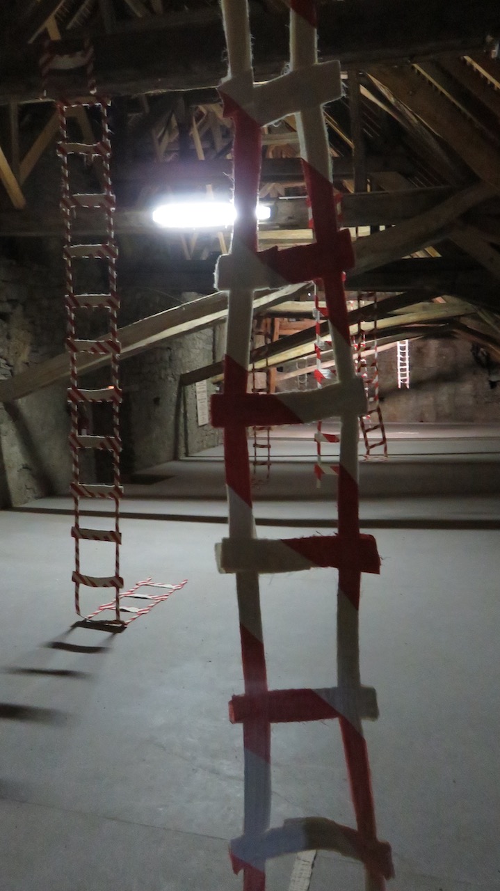 Vue de l'installation dans le grenier du château de Montmaur. Des échelles en tissu, striées de peinture rouge et blanc, tombent depuis les poutres de la charpente.