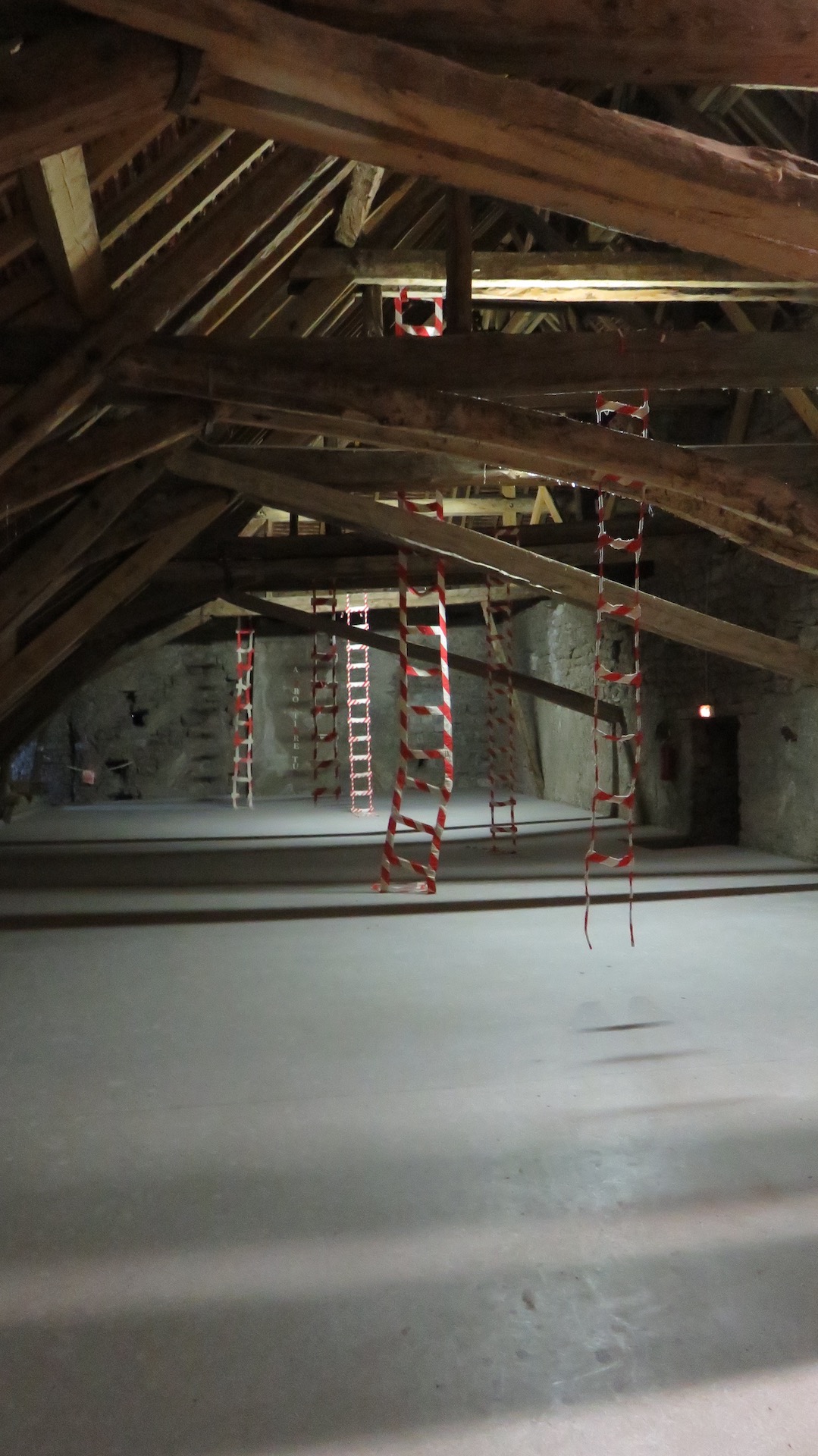 Vue de l'installation dans le grenier du château de Montmaur. Des échelles en tissu, striées de peinture rouge et blanc, tombent depuis les poutres de la charpente.