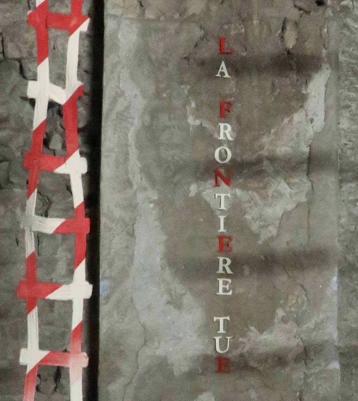 Détail de l'installation. Vue d'une échelle en tissu striée de rouge et blanc avec les mots La frontière tue sur le mur du fond