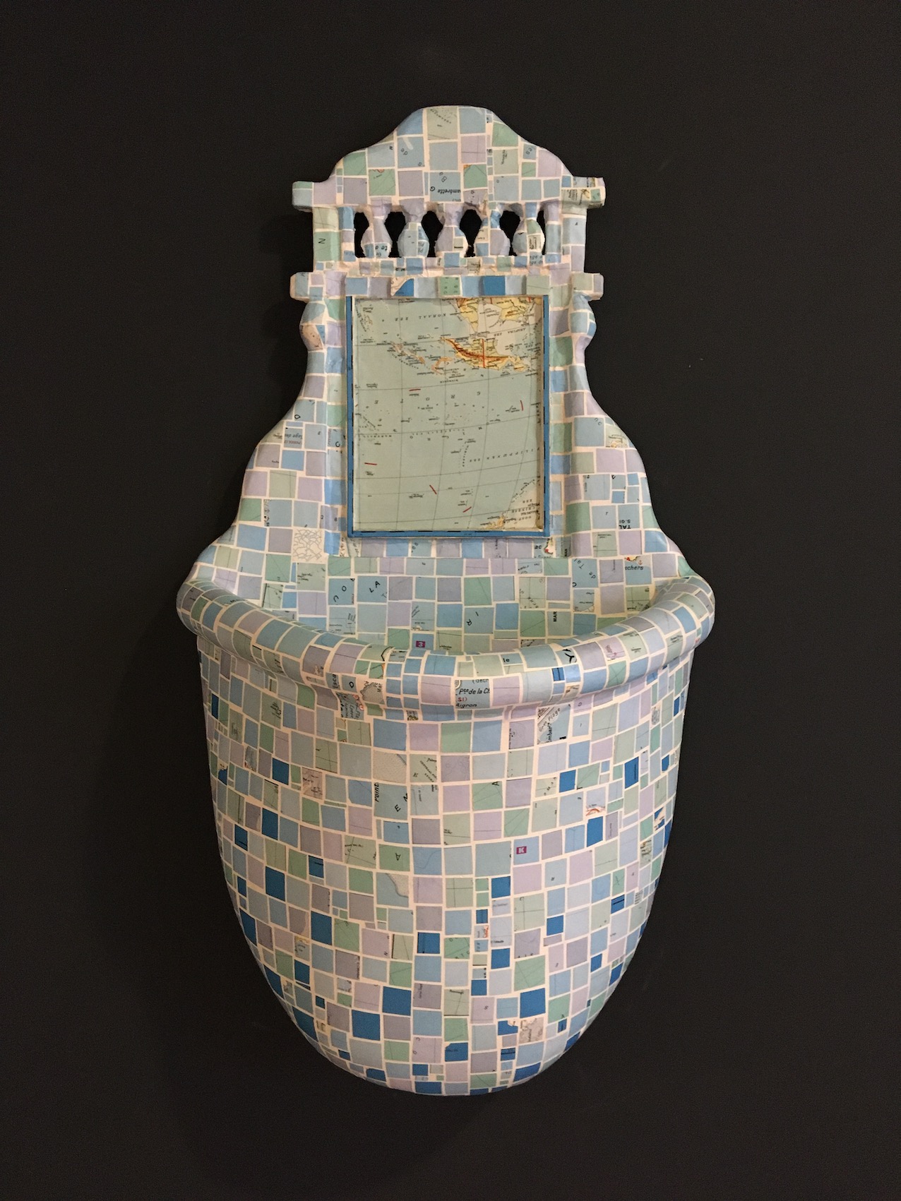 Sculpture d'un lavabo avec son miroir et le dessous du lavabo est gonflé comme une grande poche remplie. L'objet est recouvert de petits carrés de cartes de mer qui sont collés sur toute la surface comme du carrelage.