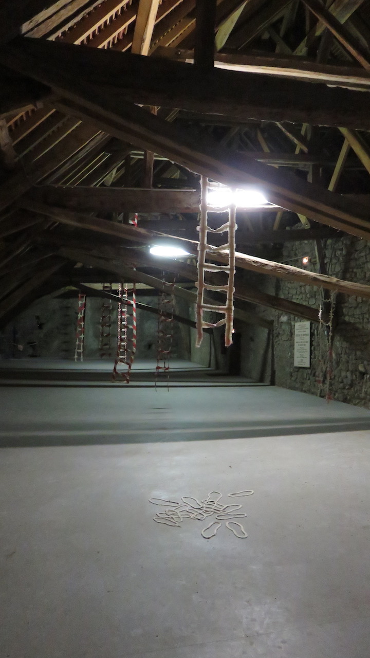 Vue d'une installation dans le grenier d'un château. Des échelles en tissu striées de rouge et blanc descendent des poutres de la charpente.