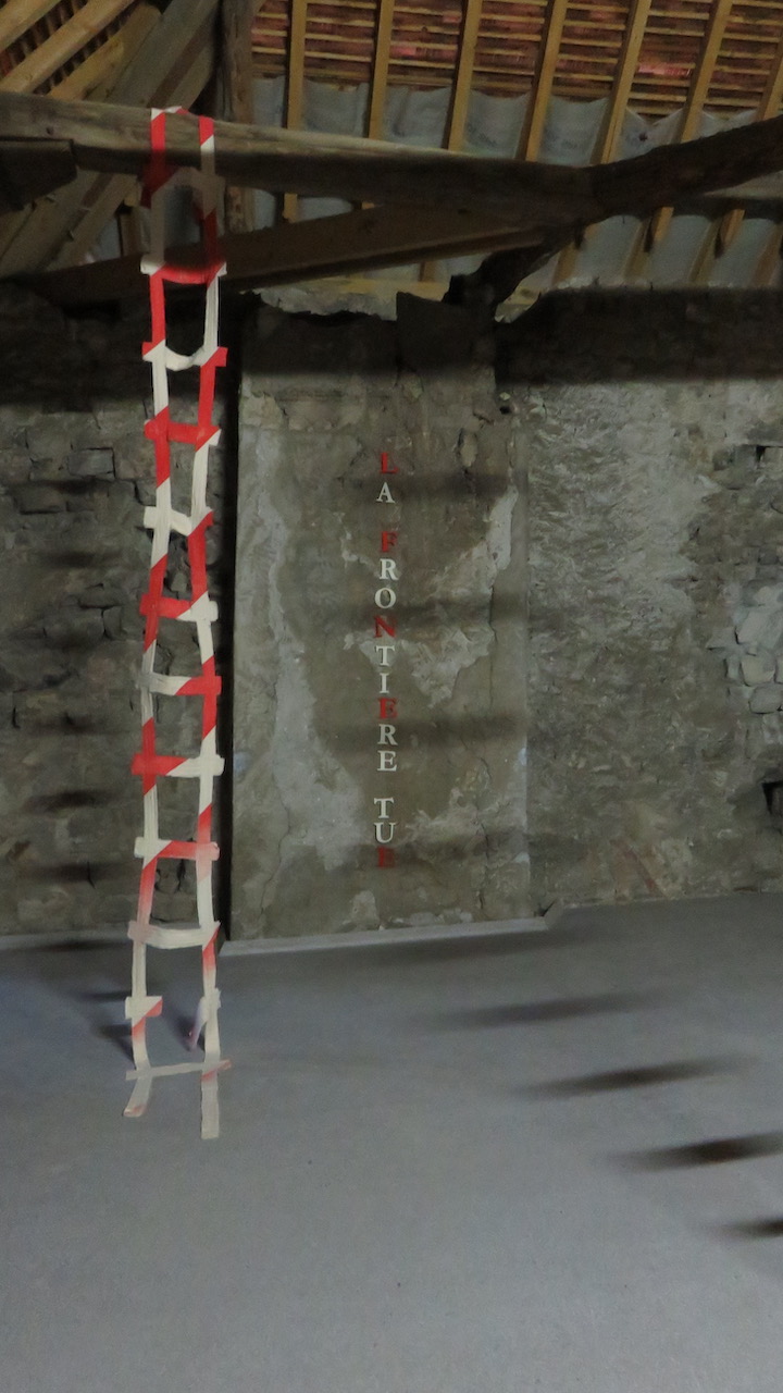 Détail de l'installation, une échelle en tissu striée de rouge et blanc descend d'une poutre, et le titre la frontière tue est accroché au mur au fond.