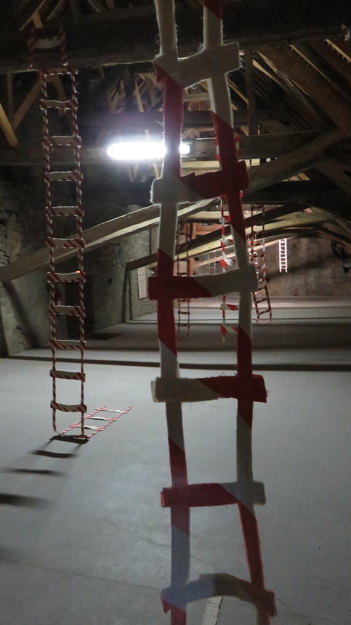 Vue d'une installation dans le grenier d'un château. Des échelles en tissu striées de rouge et blanc descendent des poutres de la charpente.