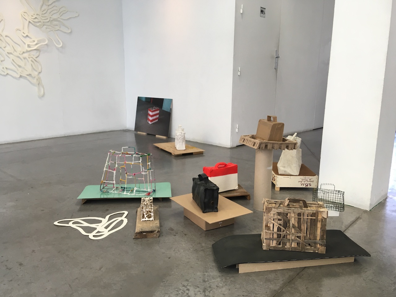 Installation de valises faites avec des matériaux variés (bois, grillage, carton, papier brulé, textile, caoutchouc,..) posées au sol sur des socles différents.