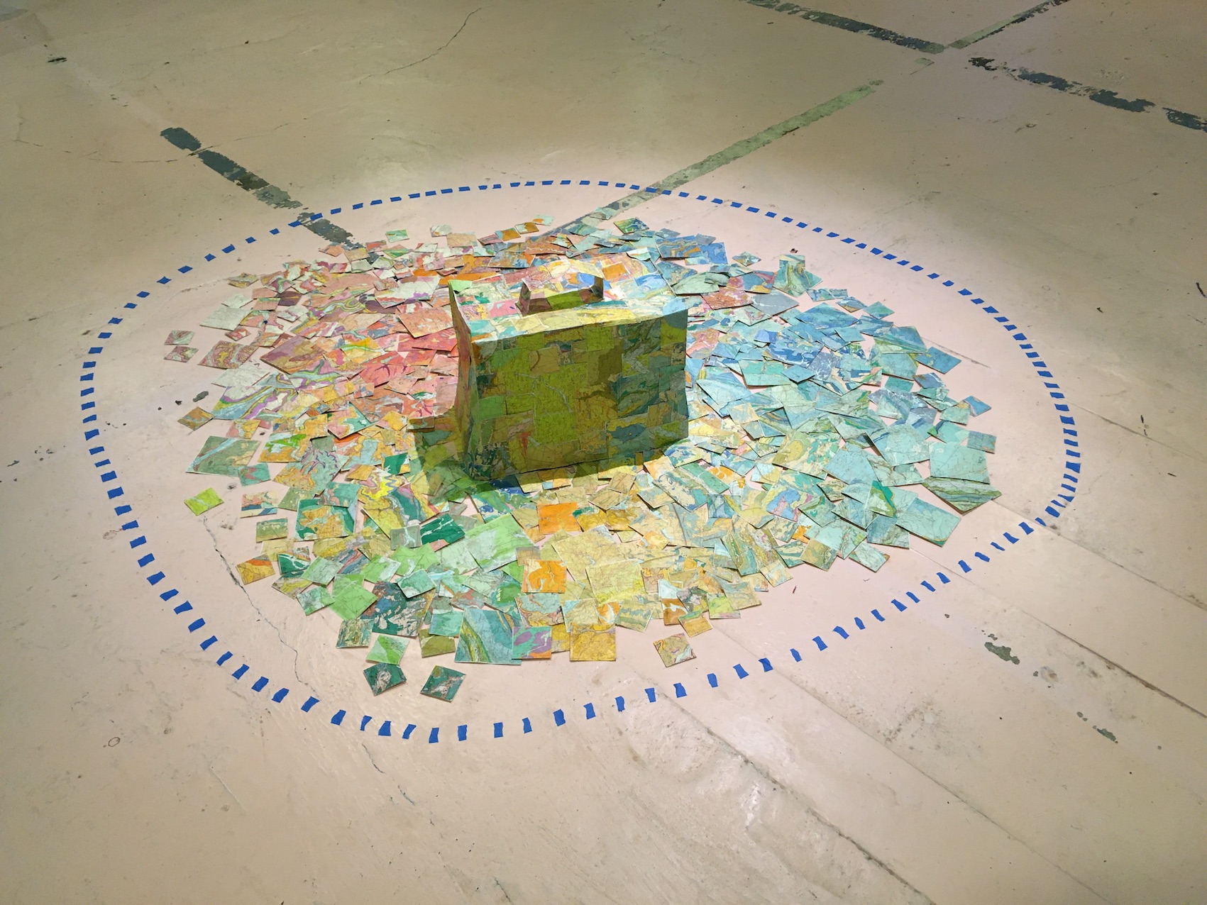 Installation au sol d'une valise faite en petits carrés de morceaux de cartes géologiques cousus les uns aux autres et qui s'étale au sol en île. Depuis l'intérieur de l'objet émane une bande son qui parle de ce que c'est 'chez-moi'