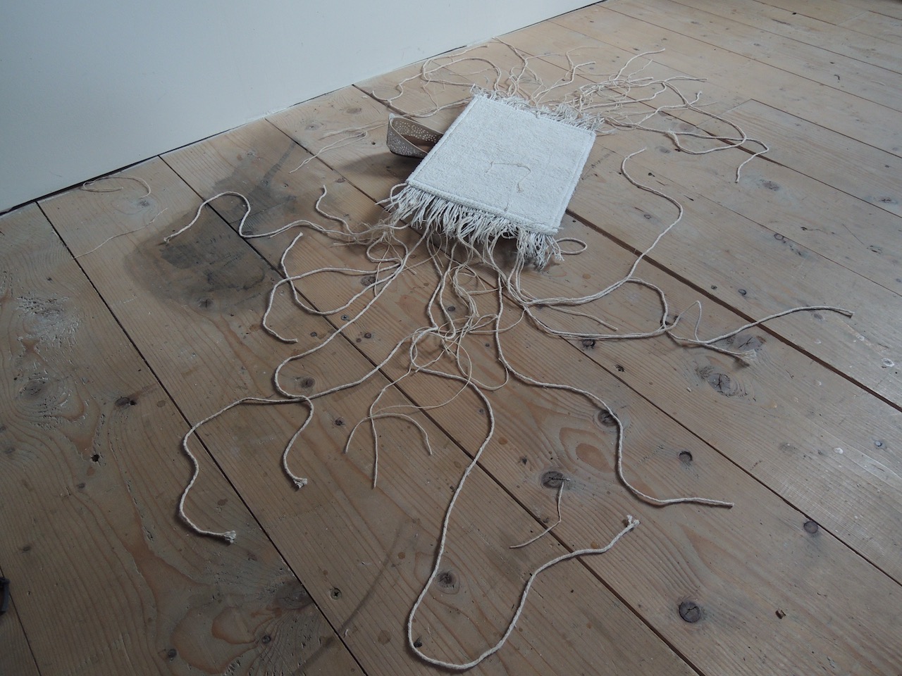 Installation au sol d'un morceau de petit escarpin blanc qui sort sous un tapis blanc dont les franges des côtés s'étalent en racines envahissantes sur le sol